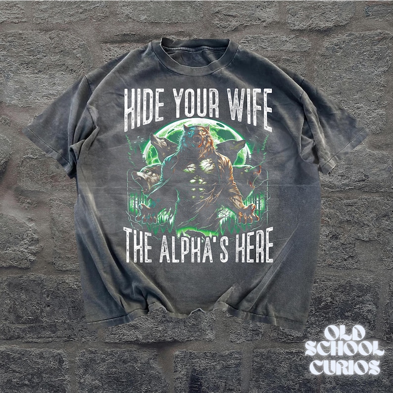 May include: Dark gray t-shirt with a werewolf graphic. The shirt features the text "HIDE YOUR WIFE THE ALPHA'S HERE" in white. The graphic includes a werewolf, wolves, and a green moon.