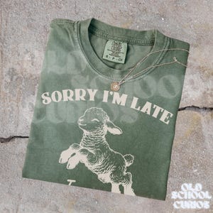 May include: A sage green t-shirt with the phrase "SORRY I'M LATE" and a graphic of a leaping lamb. A silver necklace with a small pendant rests on the shirt. The shirt has a Comfort Colors tag.