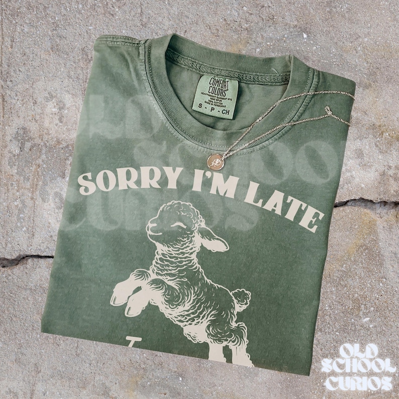 May include: A sage green t-shirt with the phrase "SORRY I'M LATE" and a graphic of a leaping lamb. A silver necklace with a small pendant rests on the shirt. The shirt has a Comfort Colors tag.