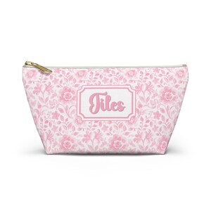 May include: A pink floral pattern cosmetic bag with a gold zipper. The bag features the name "Tites" in a pink frame. The bag is designed with a repeating pattern of pink flowers and leaves.