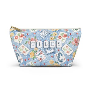 May include: A light blue zippered pouch with a floral and tile pattern. The pouch features a gold zipper and the word "TILES" in white block letters. The pattern includes red, yellow, and blue floral designs and tile illustrations.