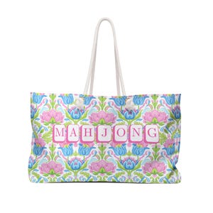 May include: A large tote bag with a white rope handle. The bag features a floral pattern in pink, blue, and green, with the word "MAHJONG" in pink letters across the front. The bag is designed for carrying items.