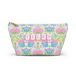 May include: A zippered pouch with a floral pattern in pink, blue, and green on a white background. The word "TILES" is printed in pink letters across the center. The zipper is gold-colored.