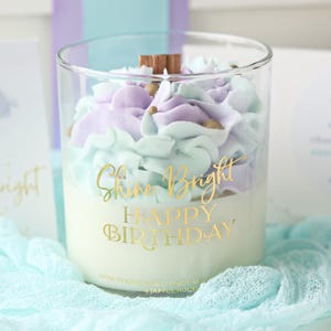 May include: A clear glass candle with a white base and pastel purple and blue frosting-like wax. The candle has gold text that reads "Shine Bright Happy Birthday". A wooden wick sits atop the wax.
