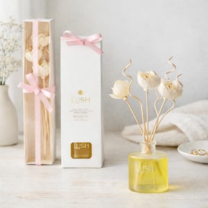 May include: Aromatherapy reed diffuser set with a clear glass bottle containing yellow liquid and white flower-shaped reeds. The set includes a white box with the Lush logo and a clear box with white flower reeds, both tied with pink ribbons.