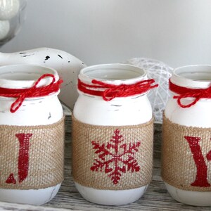 May include: Three white mason jars with red ribbon and burlap accents. The jars are decorated with red letters spelling "JOY" and a snowflake design.
