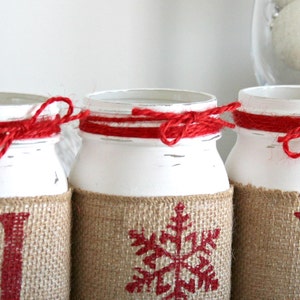 May include: Three white painted mason jars with burlap and red twine accents. The jars are decorated with red snowflake, "I", and "Y" designs.