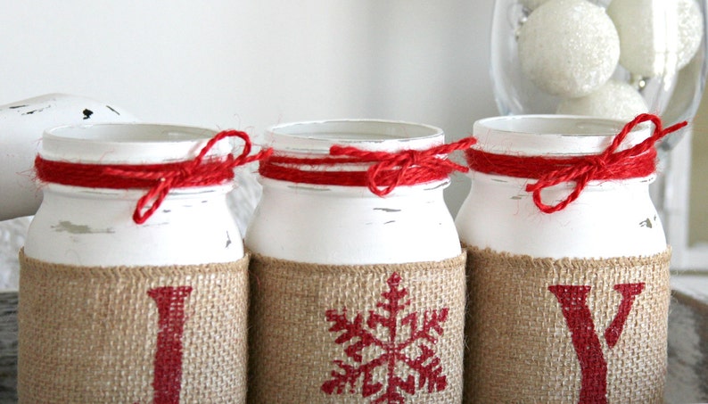 May include: Three white painted mason jars with burlap and red twine accents. The jars are decorated with red snowflake, "I", and "Y" designs.