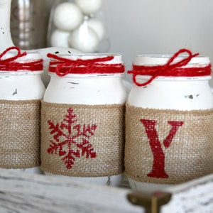 May include: Three white mason jars with red ribbon and burlap accents. The jars are decorated with the letters "J", "O", and "Y" in red paint.