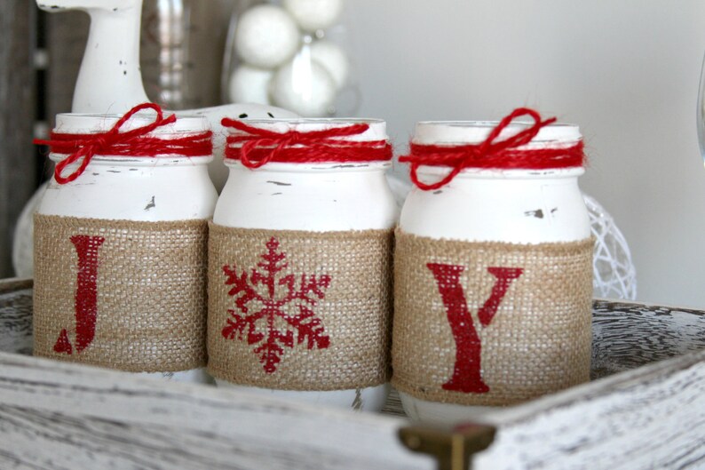 May include: Three white mason jars with red ribbon and burlap accents. The jars are decorated with the letters "J", "O", and "Y" in red paint.