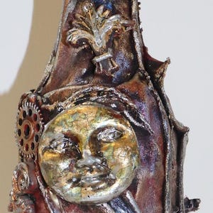 Puede incluir: Botella decorativa con temática celestial. La botella está pintada en tonos marrones, dorados y plateados, con una superficie texturizada. Presenta un diseño de sol y luna, junto con engranajes y otros adornos.