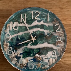 May include: Circular wall clock with a seaside theme. The clock face has a blue and green resin design with white wave accents, seashells, and a starfish. White numerals mark the hours. The clock hands are white.