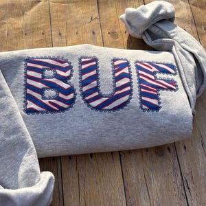 May include: A light gray long-sleeve sweatshirt featuring the letters BUF appliqued across the front. The letters are filled with a red, white, and blue zebra stripe pattern and are outlined with a dark blue decorative scalloped stitch border.