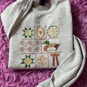 May include: A light gray crewneck sweatshirt featuring a grid of embroidered designs including green and yellow geometric quilt blocks, a brown and green mallard duck, a gold heart in an oval frame, a flower, and a brown ribbon bow.