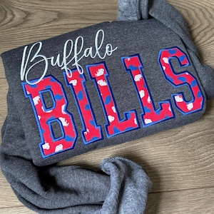 May include: A charcoal gray long-sleeve sweatshirt featuring the word Buffalo in white cursive script above the word BILLS in large block letters. The letters are red with a blue outline and a repeating pattern of small white buffalo silhouettes.