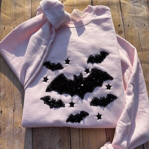 May include: A light pink crewneck sweatshirt featuring a central graphic design of five black sequined bats and several small black sequined stars scattered across the front. The garment has long sleeves and a ribbed collar with a visible white tag.