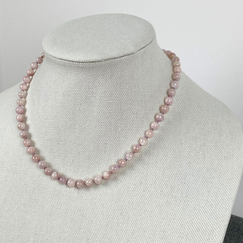 May include: A delicate necklace made of light pink and purple beads. The necklace is displayed on a white mannequin bust.