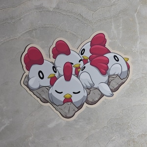 May include: A sticker featuring a group of five white cartoon chickens with red combs and yellow beaks, all nestled together in a gray textured egg carton. The chickens have simple black eyes and are depicted in various resting positions.