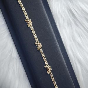 May include: A gold-coloured bracelet featuring a chain link design and three bee-shaped charms. The bracelet is presented on a black surface with a white, fluffy background. The bracelet has a lobster claw clasp.