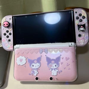 May include: A pink and purple Nintendo Switch console with a decorative case featuring cartoon characters. The case has a lace-like design and character graphics. The controllers have matching designs with character accents.