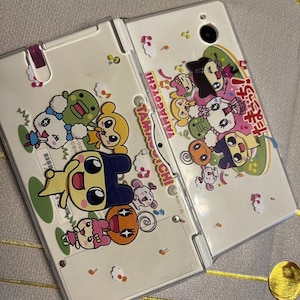 May include: A white Tamagotchi handheld game console, open and displaying colorful cartoon characters. The device features a hinged design and is decorated with various animated figures and the Tamagotchi logo.