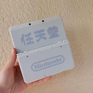 May include: A light gray Nintendo 3DS handheld game console, open to reveal the screen. The Nintendo logo is visible on the bottom half. The top half features Japanese characters. The console is held in a hand.