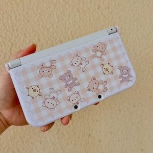 May include: A white handheld games console with a light pink gingham pattern featuring cartoon bears and chicks. The console is open, revealing the screen and buttons.