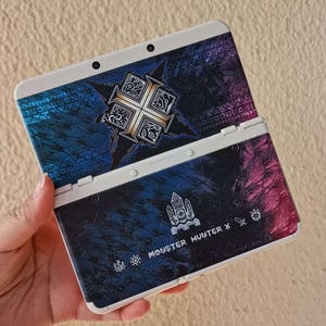 Puede incluir: Consola de juegos portátil Nintendo 3DS blanca y azul con una cubierta personalizada. El diseño es azul oscuro y morado con detalles dorados. El texto "MONSTER HUNTER X" es visible.