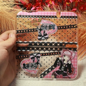 May include: Transparent phone case with a pink and black design. The case features a pink and white plaid pattern, black lace trim, and a cartoon character illustration. The case has Japanese text and a heart-shaped graphic.