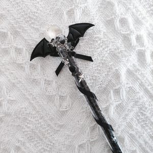 Peut inclure: Une baguette noire et argentée avec un motif d'ailes de chauve-souris. La baguette présente un globe transparent au sommet, un motif d'ailes de chauve-souris et est ornée de rubans noirs et d'accents argentés. La baguette est décorée de petits charmes argentés.