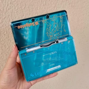 Könnte beinhalten: Türkisfarbene Nintendo 3DS-Handheld-Spielkonsole mit einer transparenten Schutzhülle. Die obere Abdeckung zeigt den goldenen Text "Solgaleo" und eine goldene Illustration. Die untere Abdeckung trägt den Text "LUNALA" und "NINTENDO 3DS".