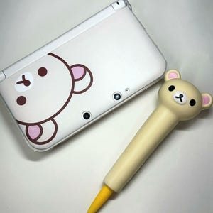 Puede incluir: Una consola de juegos portátil Nintendo 3DS blanca con un diseño de oso y un lápiz óptico a juego. El lápiz óptico es de color amarillo claro con una cabeza de oso y una punta amarilla. La consola tiene un gráfico de oso marrón y rosa.