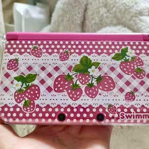 May include: Pink Nintendo 3DS case with a strawberry design. The case has a pink and white checkered pattern, strawberries, green leaves, and white flowers. The word "Swimmer" is printed on the bottom right corner.