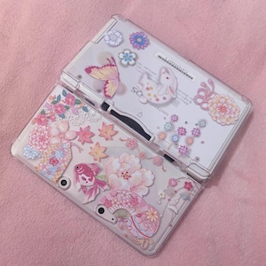 Peut inclure: Une console Nintendo DS Lite transparente avec une coque décorative ornée de motifs floraux et de papillons roses, blancs et dorés. La coque comprend un papillon rose, des fleurs et un lapin.