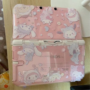 May include: A pink Nintendo 3DS LL handheld games console with a clear cover. The cover and console are decorated with cartoon character graphics in white and light purple. The console has a white hinge and buttons.