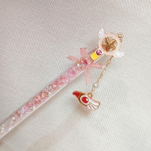 Può includere: Una bacchetta rosa e oro con un ciondolo a forma di stella e un ciondolo ad ala rosso e bianco. La bacchetta è riempita di glitter rosa e ha un fiocco rosa. È un oggetto decorativo.