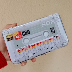 Puede incluir: Una funda transparente para teléfono diseñada para parecerse a una cinta de cassette Sony C60 vintage. La funda presenta el logotipo de Sony, el texto "Hi-Res Audio" y la palabra "Walkman" en una fuente retro. La funda es gris, roja y blanca.