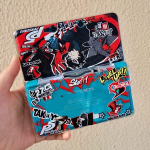 May include: A Nintendo 3DS handheld game console with a clear protective case. The case features a red, black, and white graphic design with Japanese text and the words "PERSONA 5" and "JOKER". The console is teal.