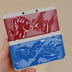 Könnte beinhalten: Zwei Nintendo 3DS Handheld-Spielkonsolen. Eine ist rot mit weißem Oberteil und einer Grafik einer Kreatur. Die andere ist blau mit weißem Oberteil und einer Grafik einer Kreatur. Auf der Oberseite jeder Konsole steht das Wort "Pokemon".