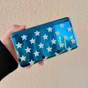 May include: A blue handheld gaming console with a white star pattern. The console is held in a clear protective case. The device has two small camera lenses and a power button.