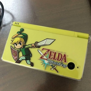 Puede incluir: Una consola de juegos portátil Nintendo DS Lite amarilla con un gráfico de Link de The Legend of Zelda: The Minish Cap. La consola tiene una cubierta protectora transparente y una lente de cámara negra.