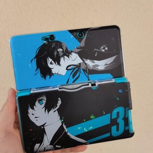 Puede incluir: Una consola de juegos portátil con una funda personalizada con ilustraciones de personajes de estilo anime. La funda tiene una combinación de colores azul y negro con un retrato de un personaje con ojos azules. También es visible el número "3".
