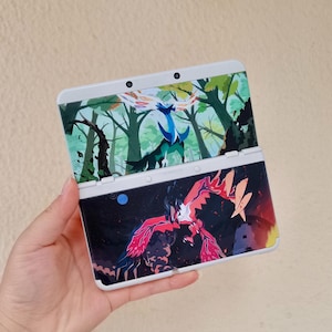 以下が含まれることがあります： 白とグレーの携帯ゲーム機。カラフルなデザイン。上画面には青と黒の生き物がいる森のシーンが表示されています。下画面には、暗い背景に赤とピンクの生き物が表示されています。