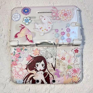 Può includere: Console Nintendo 3DS bianca con una cover protettiva trasparente. La cover presenta motivi floreali e personaggi in stile anime in rosa, bianco e viola. Sono raffigurati anche una farfalla e un coniglio.