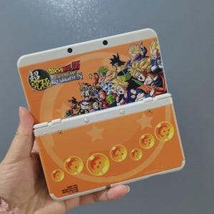 May include: Orange and white Nintendo 3DS handheld game console with Dragon Ball Z characters. The console has an orange cover with character illustrations and Dragon Balls. The text "Dragon Ball Z Extreme Butoden" is displayed.