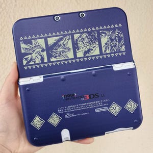 May include: A navy blue Nintendo 3DS LL handheld game console. The top cover features green illustrations of creatures and a decorative border. The console has the text "new Nintendo 3DS LL" and other Japanese text.
