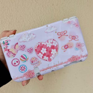 Puede incluir: Una funda de consola de juegos portátil de color lila claro con un diseño en forma de corazón lleno de flores de cerezo blancas. La funda está adornada con mariposas, nubes, lazos y elementos florales en tonos rosas y rojos. Tiene una bisagra y botones blancos.