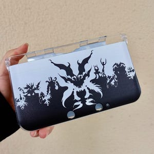 May include: A black and white Nintendo 3DS case featuring a stylized graphic design. The case has a clear plastic top and a black and white design with a detailed silhouette of figures and shapes.