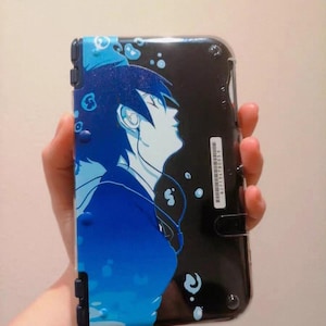 May include: A black and blue handheld gaming device case with an anime-style illustration of a person with blue hair and headphones. The design includes blue water droplet accents. The case has a clear protective cover.