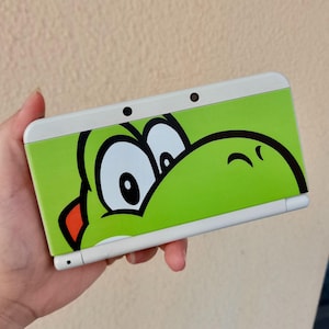 Może przedstawiać: Biało-zielona konsola do gier przenośna Nintendo 3DS z zielonym wzorem postaci Yoshi. Wzór pokrywa górną część konsoli, z wyraźnie widocznymi oczami i twarzą postaci. Konsola posiada dwa małe czarne obiektywy aparatu.